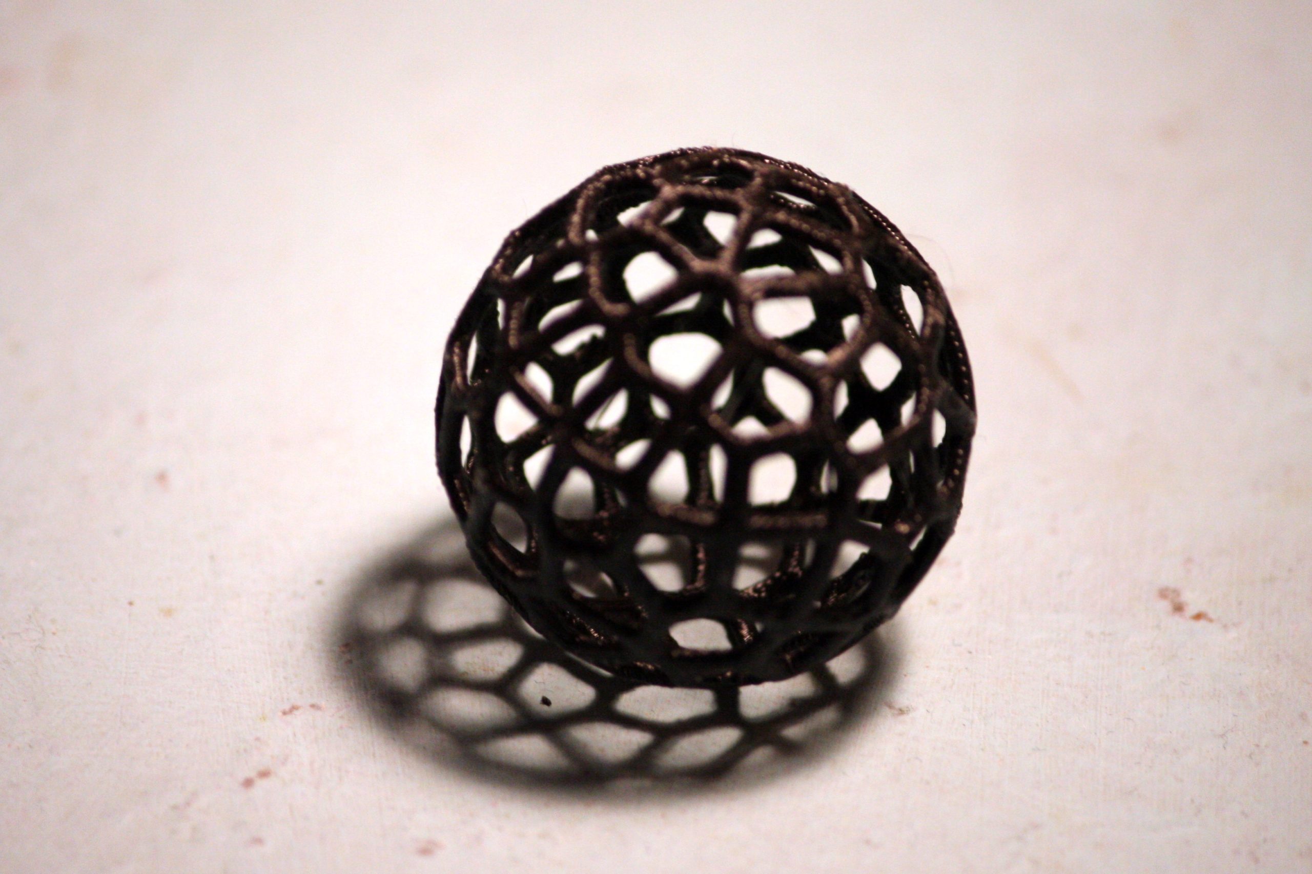 Dark 3D-printed lattice sphere casting a patterned shadow on an off-white surface; third-party illustrative reference.
